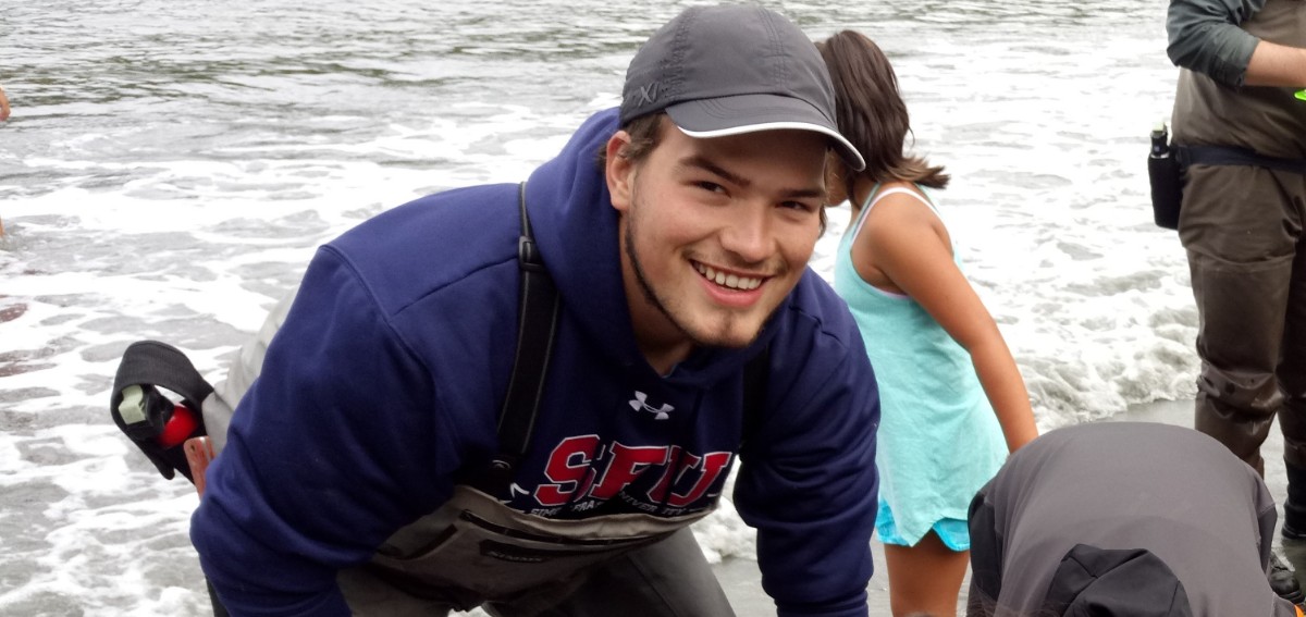Michael has a dark brown beard, and is wearing a grey cap and blue SFU hoodie. He's bending forwards and seems to be working with other people who are crouched or standing on the beach around him. He is standing on the sand and the ocean is visible directly behind him. He is smiling widely.