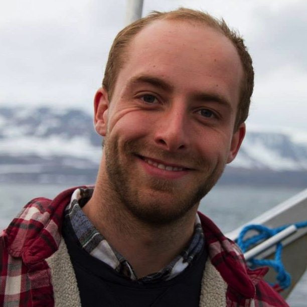A headshot of Jean who has blonde hair and beard, a red plaid shirt, and is smiling with his teeth showing. The background is a grey sky and snowy mountains. The metal rim and ropes directly behind him suggest he's on a boat.
